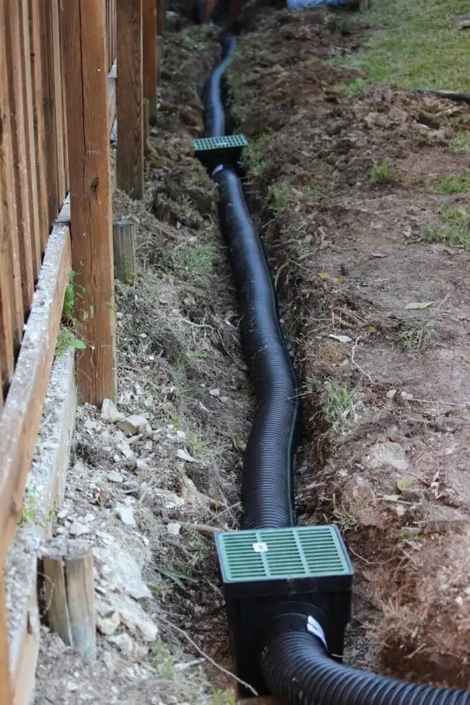 French drain installation in progress alongside a fence line — perforated corrugated pipe laid in a trench before backfilling with gravel