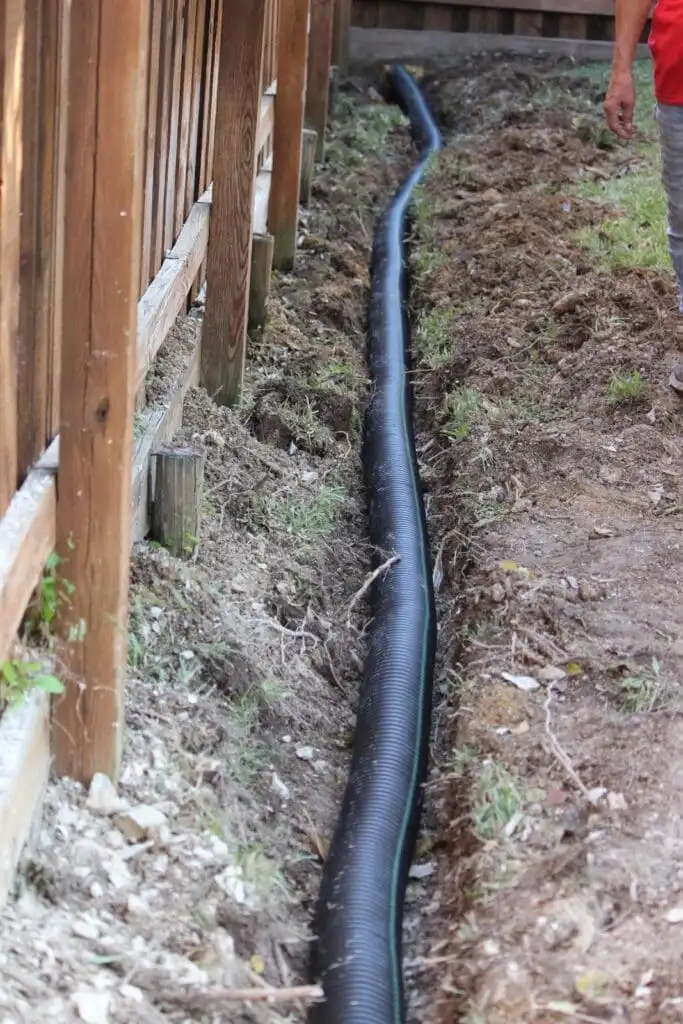 Perforated corrugated pipe laid in a trench along a fence line before backfilling with gravel, perforations facing downward