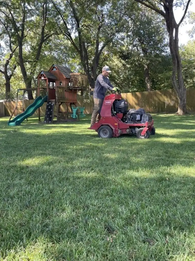 Complete Lawn Care crew member aerating a Tulsa backyard with commercial stand-on Toro aerator in fall with post oak and pecan trees overhead