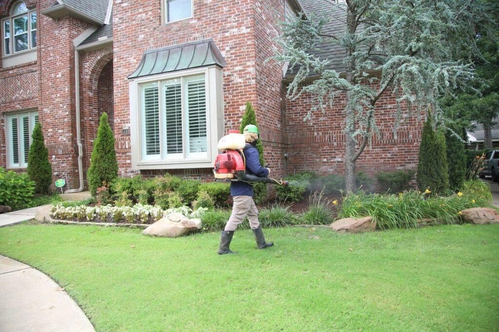 Complete Lawn Care mosquito control technician with backpack mister sprayer treating landscaping beds and shrubs with visible mist at residential property in Tulsa Oklahoma