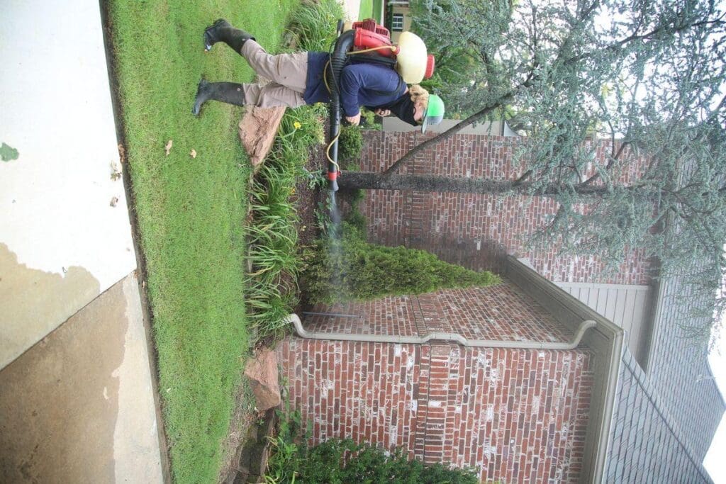 Complete Lawn Care mosquito control technician with backpack mister treating corner of brick home foundation area and landscaping in Tulsa Oklahoma
