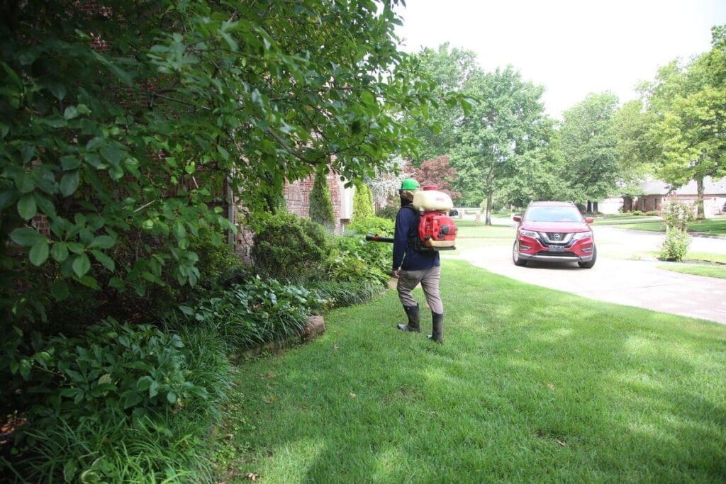 Image of Complete Lawn Care mosquito control technician with backpack sprayer treating dense vegetation and shrub line along property edge at Tulsa Oklahoma residential neighborhood