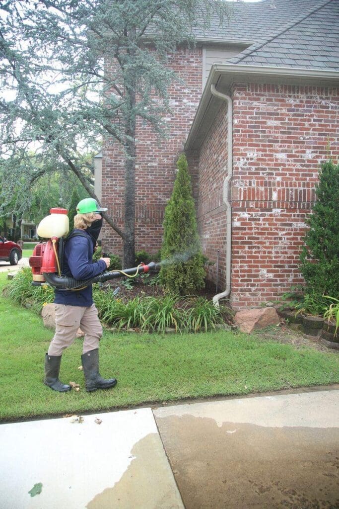 Image of Complete Lawn Care mosquito control technician with backpack mister spraying barrier treatment on foundation landscaping and shrubs at brick home in Tulsa Oklahoma