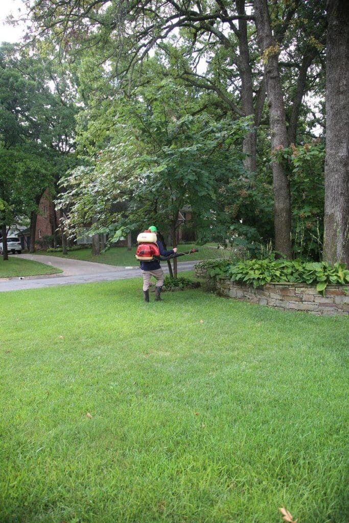 Image of Complete Lawn Care mosquito control technician with backpack sprayer treating mature trees stone retaining wall and ground cover beds at shaded Tulsa Oklahoma residential property