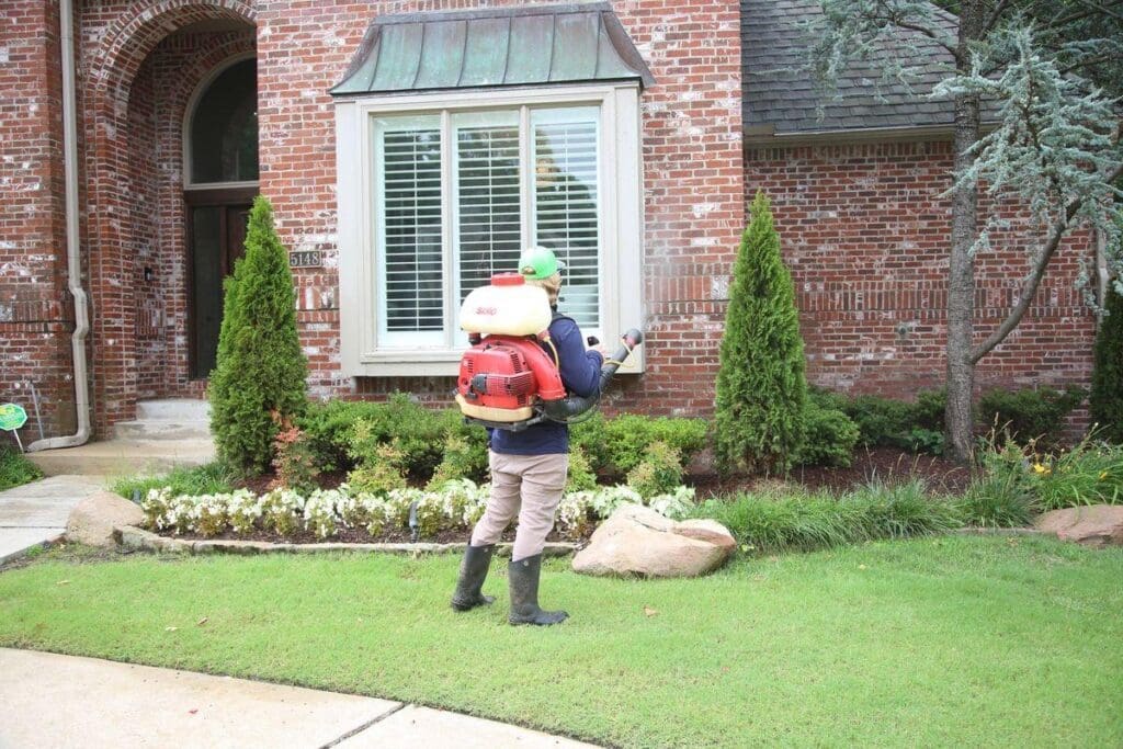Image of Complete Lawn Care mosquito control technician with backpack sprayer treating front entry landscaping flower beds and shrubs at brick home in Tulsa Oklahoma